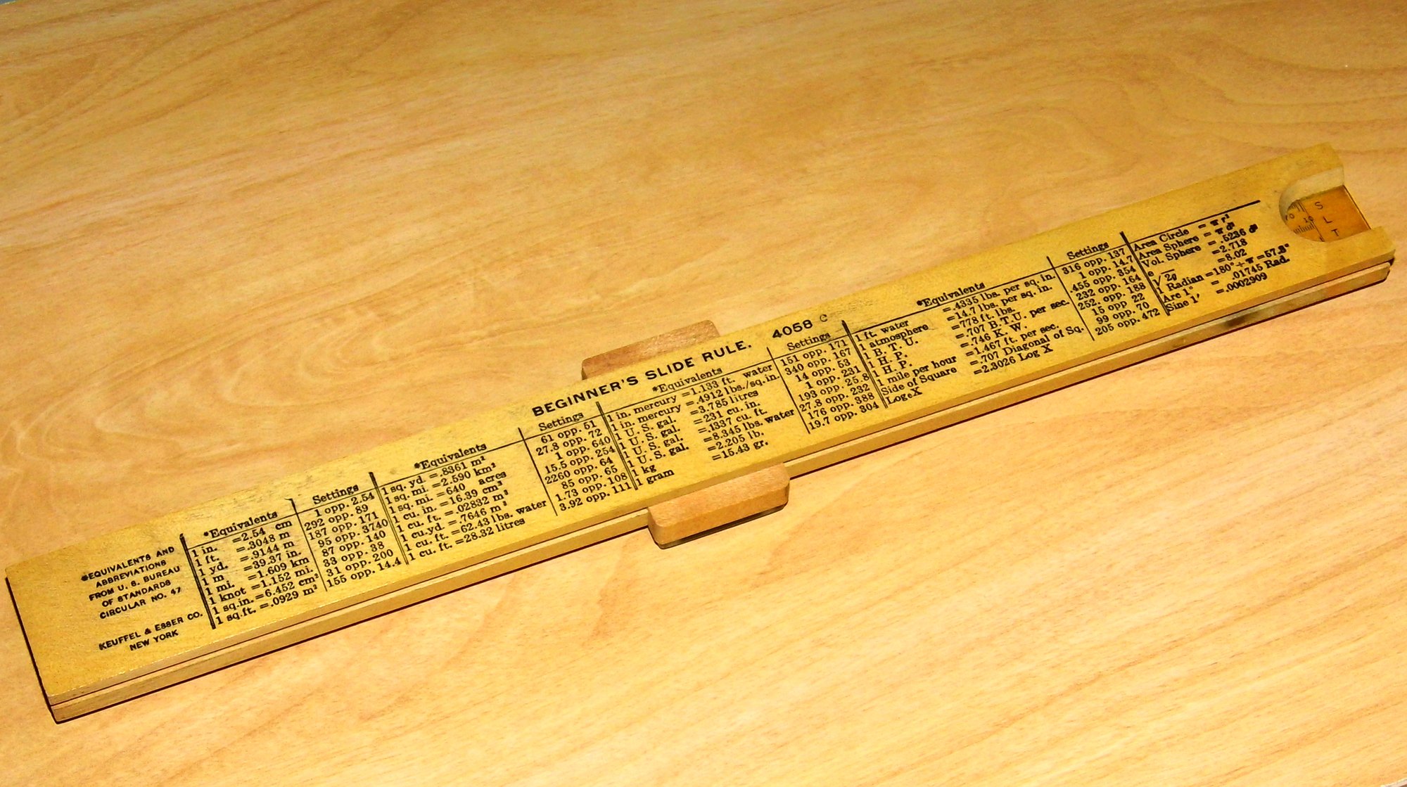 Figure 5 — The back of another K&E 4058C Beginner's Slide Rule photographed at an angle on a wooden table, with the same tables but the Bureau of Standards credit placed at the left end, and the wooden cursor…