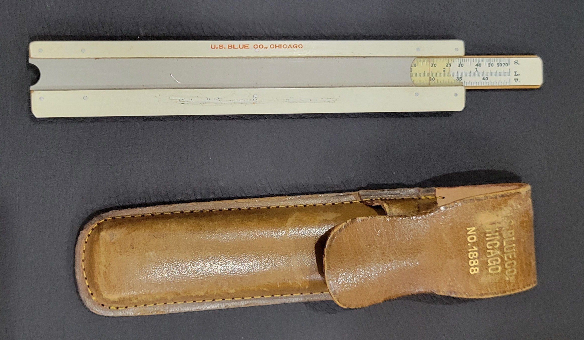 Figure 3 — The back of the U.S. Blue Co. pocket rule, printed U.S. BLUE CO., CHICAGO in red, with a grey metal backing plate, the slide drawn out to the right showing the S, L and T scales, a window at the ri…