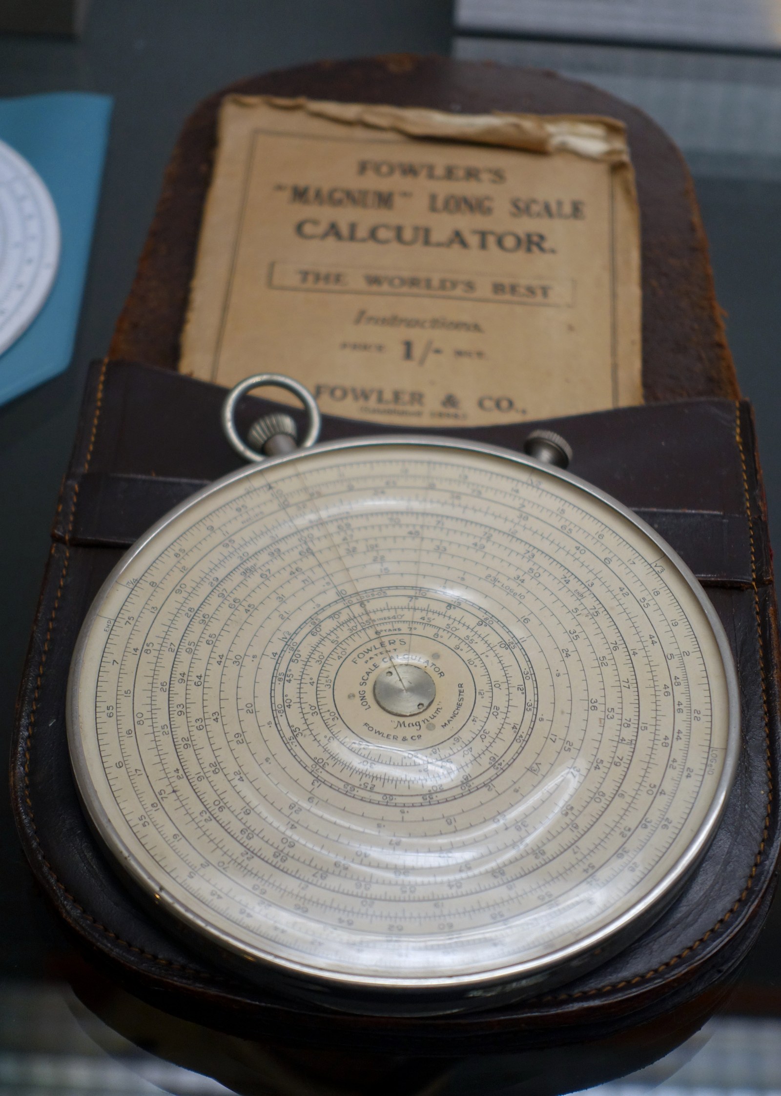 Figure 3 — A Fowler Magnum Long Scale Calculator sitting in its brown leather case, with the original paper sleeve marked "Fowler's 'Magnum' Long Scale Calculator, the World's Best" tucked behind.