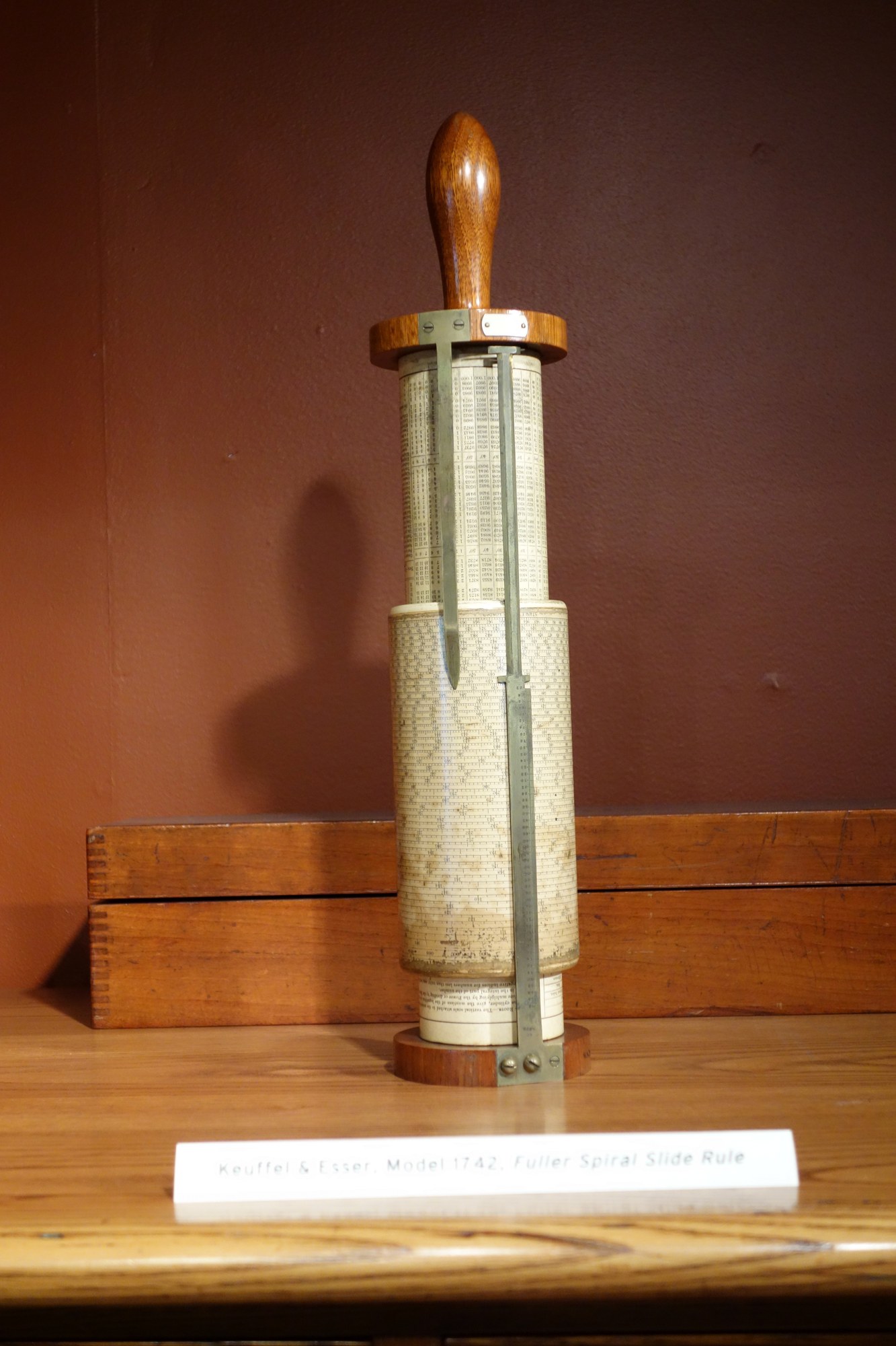 Figure 3 — A Fuller calculator standing upright on its base in a museum case, its varnished scale cylinder and brass index bar visible, labelled "Keuffel & Esser Model 1742, Fuller Spiral Slide Rule."