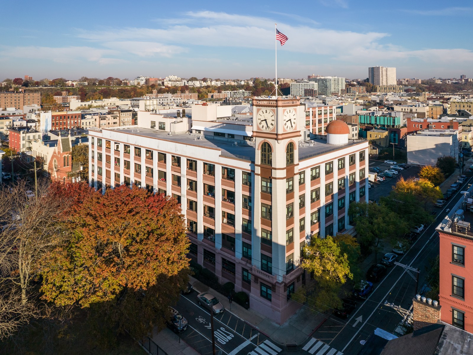 Figure 5 — An aerial view of the former Keuffel & Esser plant in Hoboken: a white-and-red five-storey factory building with a clock tower and a flag, among autumn trees and city streets.