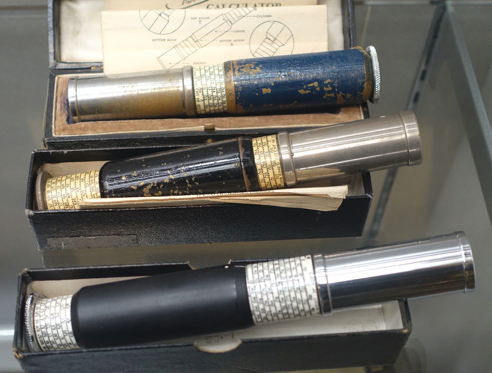 Figure 3 — Three Otis King calculators in their boxes in a museum case: a blue-and-gold early example at top with an instruction sheet, a worn black one in the middle, and a clean black-and-chrome late exampl…