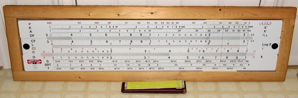Figure 3 — A large white-and-black Koh-I-Noor Graphoplex classroom slide rule, 4 ft 4 in long, lying in a plain wooden frame that formed part of its shipping carton, with a twelve-inch Pickett rule placed in …