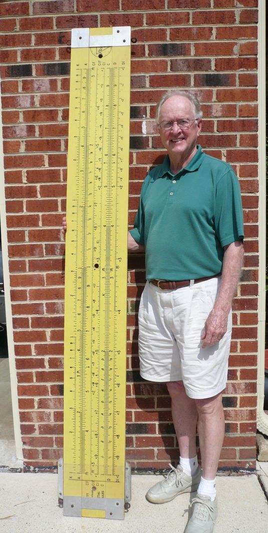 Figure 3 — A man standing beside a seven-foot yellow Pickett N1010-ES demonstration rule propped upright against a brick wall; the rule is taller than he is.